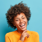 A young woman with curly hair smiles and touches her chin while wearing a bright orange sweater, standing in front of a light blue background, looking ready to call a plumber in Tampa for her next plumbing project.