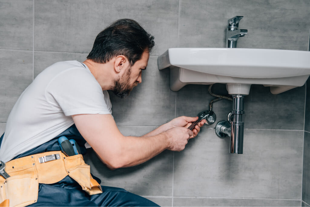 A Tampa plumber in a white shirt and tool belt uses a wrench to fix pipes under a white wall-mounted sink in a tiled bathroom, showcasing expert plumbing skills.