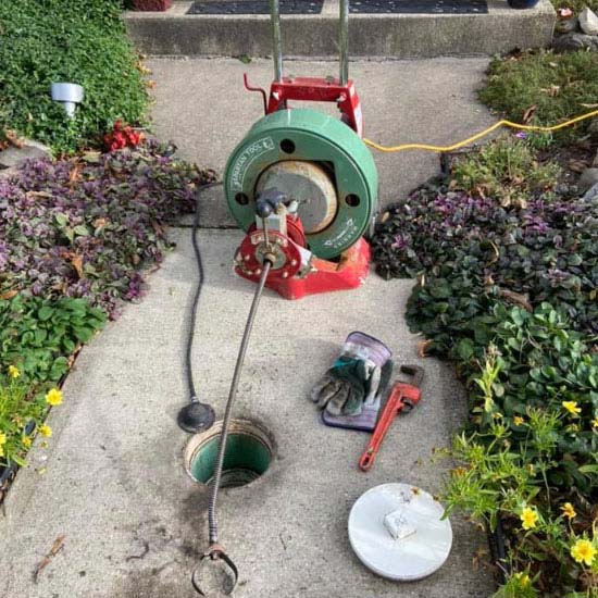 A Tampa plumber sets up a drain snake machine on the sidewalk, its cable inserted into an open sewer access hole, surrounded by gloves, a pipe wrench, and a removed cover.
