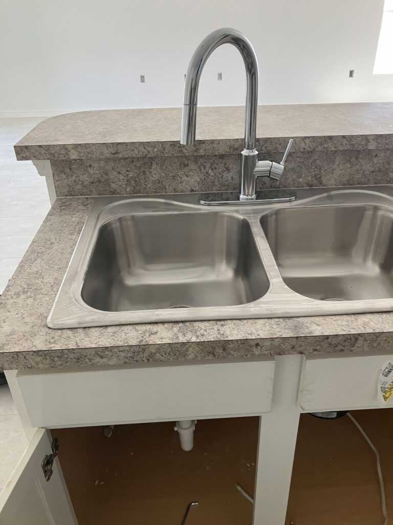 A double-bowl stainless steel kitchen sink with a tall faucet installed in a speckled countertop; cabinet doors below are open, revealing plumbing underneath—perfect for inspection by your trusted Tampa plumber.
